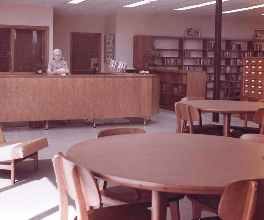 A librarian sitting at the checkout desk reading through library cards circa 1960.