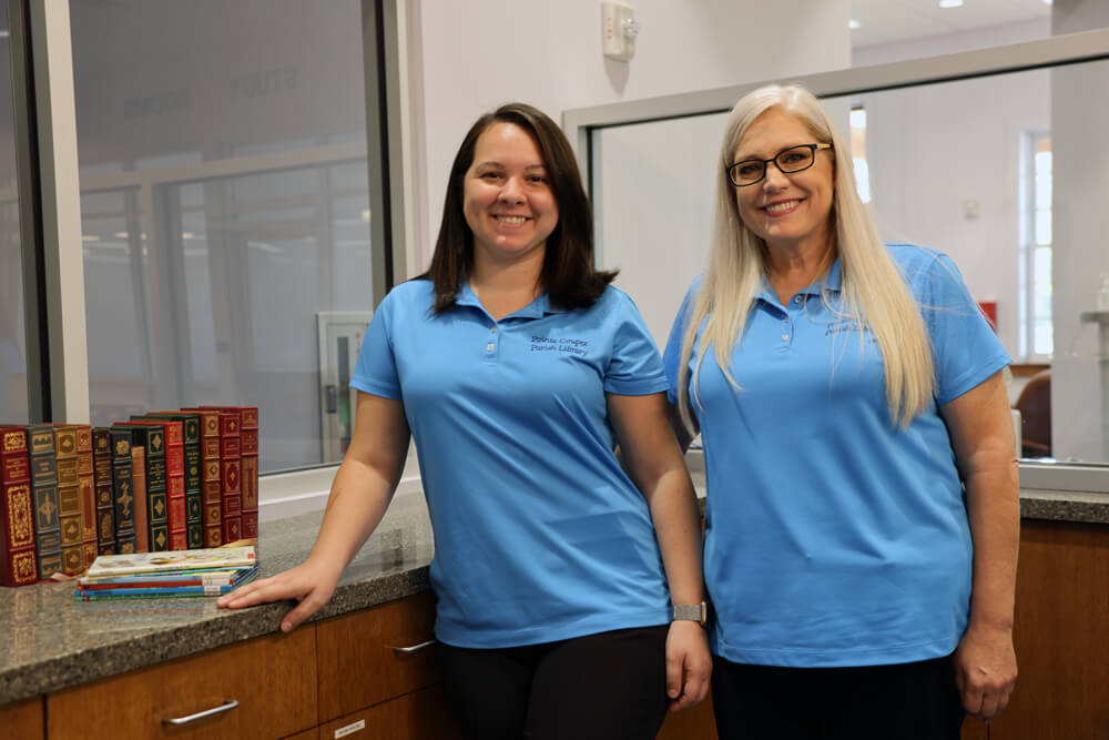 Two Pointe Coupee library staff members in periwinkle clothing smiling at the receptionist's desk.