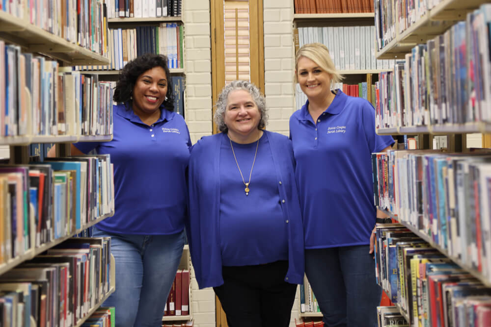 Three Pointe Coupee Library staff members in indigo tops smiling down a row of bookshelves.
